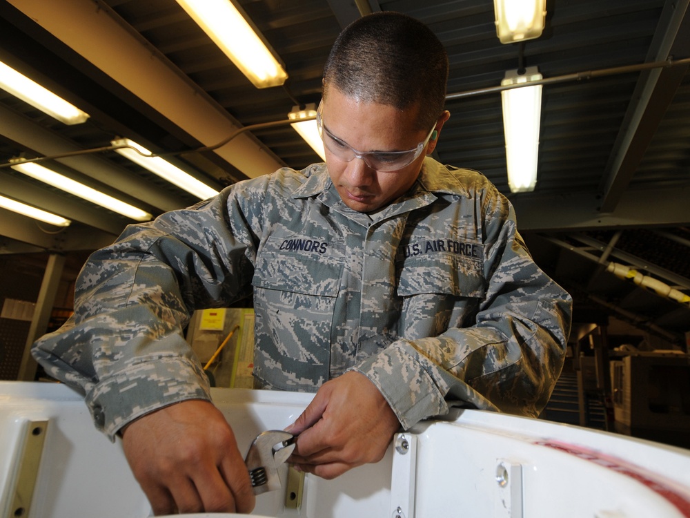 A U.S. Air Force service member named Connors is performing maintenance or repair work on what appears to be an aircraft component or panel. The airman is wearing the standard Airman Battle Uniform (ABU) in digital tiger-stripe camouflage pattern along with safety glasses. The work is being conducted indoors in what appears to be a maintenance hangar with fluorescent overhead lighting.