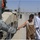 A U.S. soldier in ACU (Army Combat Uniform) is handing a small item, likely candy or a snack, to a local child on a road in what appears to be Iraq or Afghanistan. Three children are visible, with two boys standing nearby watching the exchange. A convoy of military HMMWV (Humvee) vehicles is parked along the road, with a STOP sign visible on the lead vehicle.