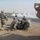 The image shows multiple soldiers in U.S. Army ACU (Army Combat Uniform) pattern camouflage attending to a downed individual on a dirt road. Smoke or dust is visible in the background, suggesting a recent incident. A military vehicle, likely a HMMWV (Humvee), is partially visible on the right side of the frame.