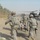 Military personnel in U.S. Army combat uniforms are carrying a casualty on a litter toward a waiting helicopter, consistent with a MEDEVAC operation. The scene appears to take place on or near a road in an arid environment with palm trees visible in the background. A UH-60 Black Hawk helicopter is on the ground with rotors spinning, ready for immediate departure.