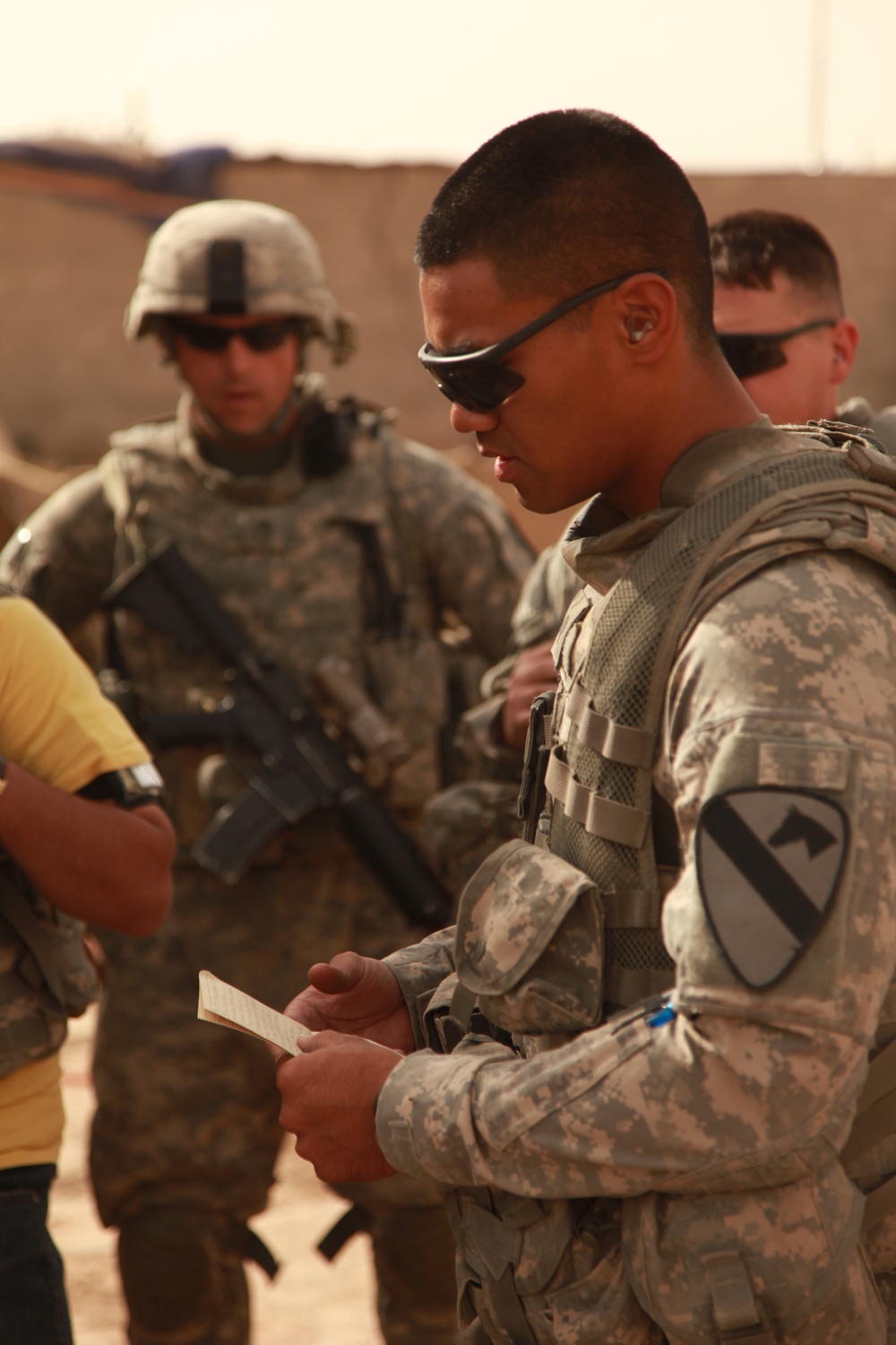 The image shows U.S. Army soldiers in a desert environment, wearing Army Combat Uniforms (ACU) with full gear. The foreground soldier is wearing a 1st Cavalry Division patch and is reviewing a small document or notecard. The background shows at least two other soldiers, one wearing a combat helmet and carrying an assault rifle.