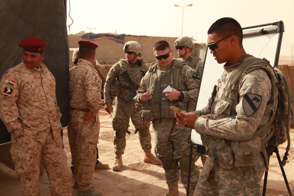 The image shows a joint military meeting or briefing between U.S. Army soldiers and what appear to be Iraqi military personnel in a desert environment. U.S. soldiers in ACU (Army Combat Uniform) pattern camouflage are gathered near a whiteboard, reviewing documents and notes. Two personnel in desert camouflage uniforms with red berets, bearing Iraqi military insignia, are present on the left side.