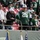 New York Jets Vs Jacksonville Jaguars Military Ceremony A New York Jets NFL game featuring fans in the stands and a military color guard ceremony on the field level. A prominent fan wearing a Jets #25 jersey, hard hat, and green Hulk hands stands at a railing with two children in Jets gear. Below, uniformed military personnel carry flags including the US flag and service branch flags.