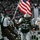 New York Jets Vs Jacksonville Jaguars Military Ceremony New York Jets NFL players are entering a stadium field, with one player carrying a large American flag. The team is dressed in their green and white home uniforms, with visible jersey numbers and names including Revis (#24) and number 25. A large crowd of spectators fills the stadium stands in the background.