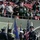 New York Jets Vs Jacksonville Jaguars Military Ceremony A military color guard ceremony is taking place on the field of a New York Jets NFL game. Members of multiple military branches — including the Air Force, Army, and Marine Corps — are standing at attention holding flags, including the American flag and service branch flags. Fans in New York Jets green jerseys and gear are visible in the stadium stands above.