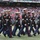 New York Jets Vs Jacksonville Jaguars Military Ceremony A formation of U.S. Marine Corps personnel in dress blue uniforms are marching on the field of a New York Jets NFL stadium. The Marines are wearing their distinctive dress blues with white covers (caps), white gloves, and red-striped trousers, indicating a formal ceremonial appearance. A stadium scoreboard in the background advertises 'The First Ever Jets Flight Crew Swimsuit Calendar,' confirming the NFL venue.
