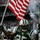New York Jets Vs Jacksonville Jaguars Military Ceremony A New York Jets NFL player wearing jersey number 25 leads teammates onto the field while carrying a large American flag. The scene appears to be a pre-game entrance ceremony, likely around 2009-2010 based on the '50th Anniversary' patch visible on the jersey and the scoreboard text reading 'BUY YOUR 2010'. Pyrotechnic smoke effects and a packed stadium crowd are visible in the background.