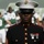 New York Jets Vs Jacksonville Jaguars Military Ceremony A United States Marine Corps staff sergeant stands at attention in dress blues uniform at what appears to be an NFL football game. The Marine is in the foreground in sharp focus, wearing the distinctive blue dress uniform with red trim, gold buttons, white cover, and service ribbons with miniature medals. In the blurred background, football players in green and white uniforms (consistent with the New York Jets) are visible.