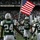 New York Jets Vs Jacksonville Jaguars Military Ceremony New York Jets NFL players are running onto the field during a pre-game ceremony at what appears to be Giants Stadium (now MetLife Stadium). A player carrying an American flag leads the team entrance, with a packed crowd of fans visible in the background. A CBS2 'New York's Sports Leader' banner is visible in the stadium, along with an NFL-vested cameraman capturing the moment.