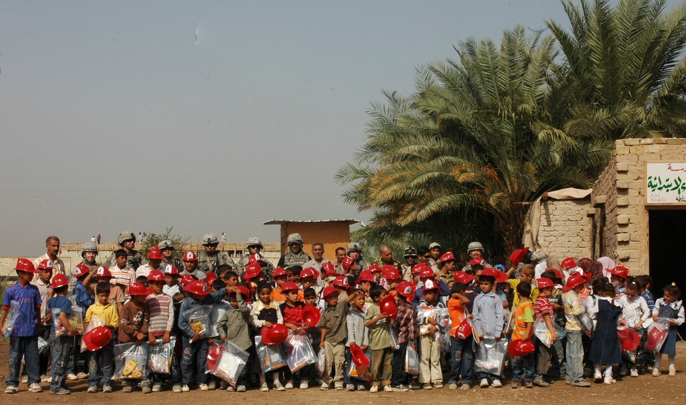 A large group of children wearing red hard hats are gathered outdoors alongside several military personnel in desert camouflage uniforms. The children appear to be holding plastic bags containing supplies or gifts. The setting appears to be a rural or semi-urban area in the Middle East, with palm trees and mud-brick structures visible in the background.