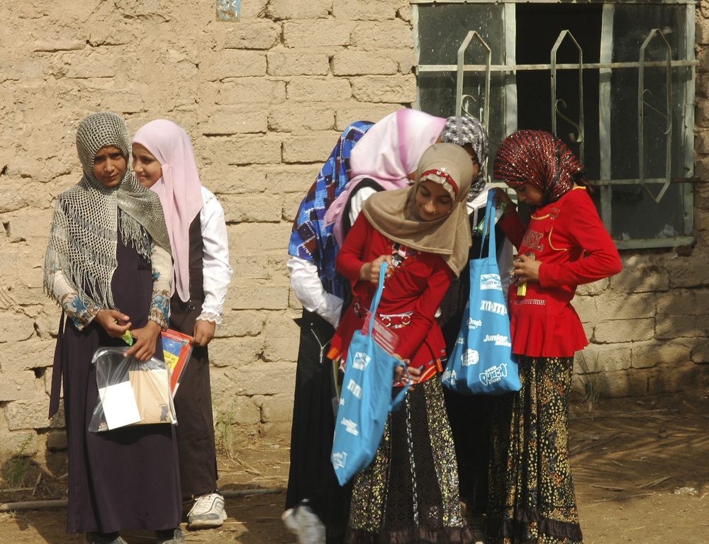 A group of girls and young women wearing hijabs are gathered outside a mud-brick building. They appear to be examining or distributing items from blue tote bags and plastic-wrapped packages. The setting appears to be a modest rural or semi-urban environment.