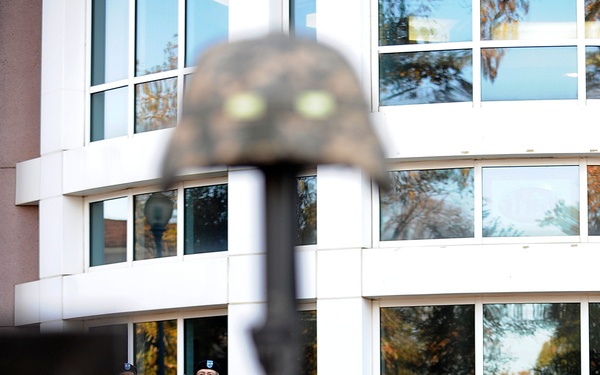 A military ceremony is taking place outdoors in front of a modern building, with a soldier reading from papers while others stand at attention in the background. The foreground features a 'Battlefield Cross' memorial — a rifle with a helmet balanced on the stock — a traditional tribute to fallen soldiers. The composition frames the ceremony through this symbolic memorial in the foreground.