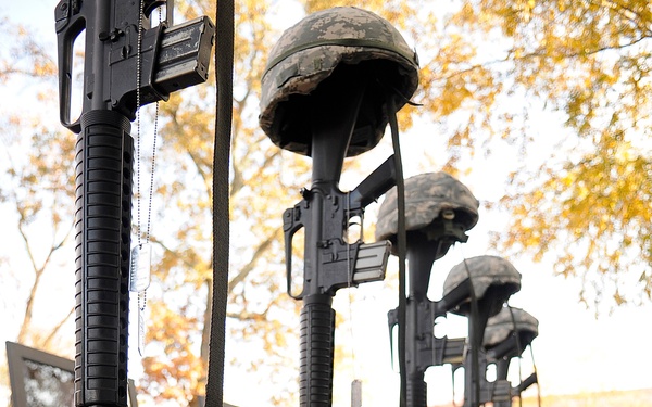 The image depicts a military memorial display, commonly known as a 'Battle Cross' or 'Fallen Soldier Battle Cross' tribute. Multiple sets of tan/desert combat boots are arranged in a row on a black-draped surface, each paired with an inverted rifle (bayonet down) topped with a combat helmet and dog tags hanging from the rifles. The display appears to honor multiple fallen soldiers, set outdoors with autumn trees visible in the background.