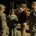 A nighttime scene at a military base showing a civilian in civilian clothing (red sweater, khaki pants, dark cap) engaged in conversation with several U.S. Army soldiers in ACU (Army Combat Uniform) camouflage. The group is standing on gravel near military vehicles, including what appears to be an MRAP (Mine-Resistant Ambush Protected) vehicle in the background. The setting suggests a VIP or dignitary visit to a forward operating base.