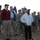 A group of civilians and U.S. Army soldiers are gathered outdoors in what appears to be an arid, rocky desert environment, likely during a military visit or congressional delegation (CODEL). Several civilians in casual attire stand among uniformed soldiers wearing ACU (Army Combat Uniform) pattern camouflage. Military unit flags/guidons are visible in the background, suggesting an official or ceremonial context.