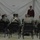 A civilian in a dark red sweater and cap appears to be addressing a group of military personnel seated in chairs arranged in a semicircle inside a large tent or temporary structure. The soldiers are wearing US Army combat uniforms (ACUs). This appears to be a briefing, presentation, or morale event at a forward operating base or military installation.