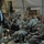 A civilian man in a dark jacket and khaki pants stands and addresses a group of military personnel seated in chairs inside what appears to be a large tent or temporary structure. The soldiers are wearing US Army combat uniforms (ACU pattern) and appear to be attentively listening to the speaker. The setting suggests a briefing, town hall meeting, or morale visit at a forward operating base or military installation.
