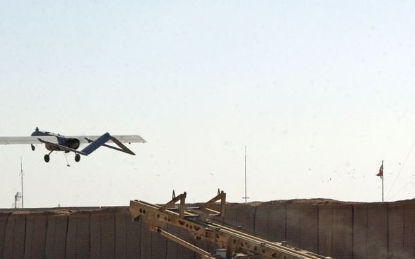 A fixed-wing unmanned aerial vehicle (UAV) is airborne immediately after being launched from a catapult launcher system in what appears to be a forward operating base in a desert environment. The launch ramp, mounted on a wheeled trailer, is angled upward and still shows signs of recent use. The surrounding area features concrete T-wall barriers, communication antennas, and arid terrain consistent with a military installation in the Middle East.