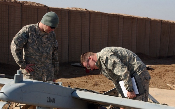 Two U.S. Army soldiers in ACU (Army Combat Uniform) camouflage are conducting a pre-flight inspection or maintenance check on a fixed-wing UAV (unmanned aerial vehicle) marked with the number '2048', consistent with an RQ-7 Shadow drone. The aircraft is mounted on a ground support launcher/cradle in what appears to be a forward operating base in an arid environment. One soldier is observing while the other leans in closely, holding a clipboard or checklist.