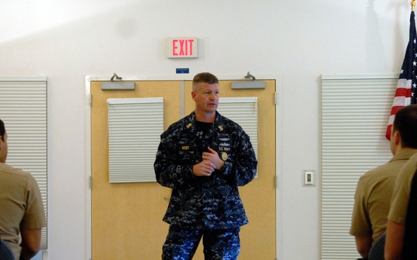 A U.S. Navy officer in Navy Working Uniform (NWU) Type I blue digital camouflage is addressing a group of seated military personnel in what appears to be a briefing or training session. The audience, wearing tan/khaki uniforms consistent with U.S. Marine Corps or Navy working attire, is seated in rows on both sides of a center aisle. The setting is an indoor facility with an American flag and a Navy flag visible in the background.