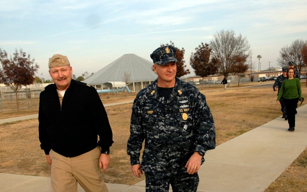 Two U.S. Navy personnel walk side by side along a concrete sidewalk on what appears to be a military installation. The individual on the right wears the Navy Working Uniform (NWU) digital camouflage pattern with a name tape reading 'WEST' and a cover, while the individual on the left wears service khakis with a garrison cap and shoulder insignia. Several civilians and additional military personnel follow behind them on the same path.
