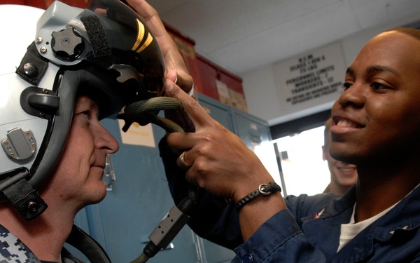Two military personnel are shown in what appears to be a locker room or equipment room. One individual wearing a Navy digital camouflage uniform is being fitted with a flight helmet by a second individual in a dark blue uniform. The person assisting is smiling while adjusting the helmet's oxygen mask or chin strap connections.