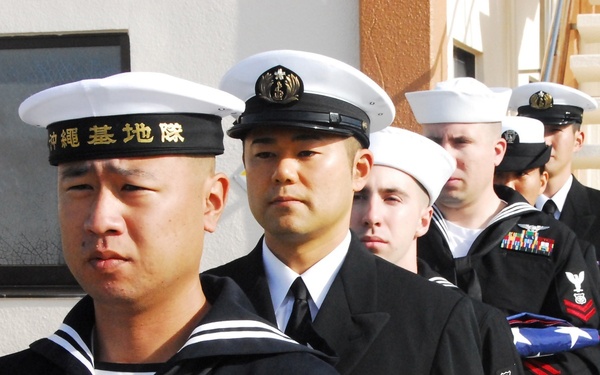 A formal military ceremony featuring both Japanese Maritime Self-Defense Force (JMSDF) and United States Navy personnel standing in formation. The personnel are in dress uniforms and are holding folded flags, suggesting a ceremonial or memorial event. The group appears to be in a joint military context, likely at a shared base in Okinawa, Japan.