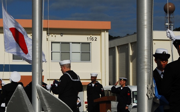 A formal military ceremony is taking place at what appears to be a U.S. Navy installation, with personnel in dress uniforms participating in a flag-raising or change-of-command ceremony. The Japanese national flag is visibly being raised on one of the flagpoles, suggesting a joint U.S.-Japan naval event. Large ship anchors and chain barriers are displayed in the foreground, consistent with a naval base setting.