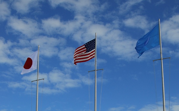 A formal military ceremony is taking place at a Port Operations facility designated 'CFAO,' with a large formation of naval personnel in dress uniforms assembled in front of three flagpoles. The flags of Japan, the United States, and what appears to be the United Nations are flying. Large ship anchors are displayed as decorative elements within a chained perimeter around the flagpole area.