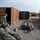 Two soldiers in US Army ACU (Army Combat Uniform) camouflage are working outdoors on a gravel surface between shipping containers at what appears to be a forward operating base or military installation. They are handling large camouflage-covered equipment bags or cases laid out on the ground. The setting includes multiple intermodal shipping containers, a portable toilet, and desert-like terrain consistent with a Middle Eastern deployment.