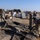 A group of U.S. Army soldiers in ACU (Army Combat Uniform) camouflage are working together at what appears to be a forward operating base or staging area in an arid desert environment. Several soldiers are handling and spreading out a large dark military tarp or tent material on the gravel ground. The background shows shipping containers, military equipment, pallets, and additional personnel engaged in various logistics tasks.