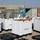 Military personnel in ACU (Army Combat Uniform) camouflage are working among numerous white reusable containers arranged on a gravel surface. The scene appears to be a forward operating base or logistics staging area, with multiple intermodal shipping containers visible in the background. Soldiers appear to be inventorying, unpacking, or organizing supplies from the white containers.