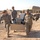 Three U.S. Army soldiers in ACU (Army Combat Uniform) camouflage are carrying a cardboard box filled with miscellaneous equipment and materials at what appears to be a forward operating base (FOB) in a desert environment. Two soldiers carry the box while a third walks alongside. The background shows typical military base infrastructure including HESCO barriers, shipping containers, and prefabricated structures.