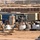 A military forward operating base (FOB) logistics area showing a busy supply and distribution yard. Multiple shipping containers in various colors are arranged throughout the compound, with personnel in military uniforms handling cargo and equipment. The background shows HESCO barriers, satellite dishes, and temporary structures typical of a deployed military installation.