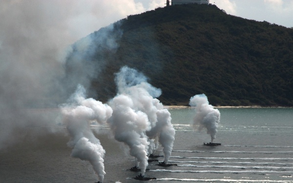 Multiple military amphibious assault vehicles are traversing a coastal bay while deploying white smoke screens, likely during a military exercise or amphibious landing drill. The vehicles are moving in a formation toward shore, leaving wake trails in the water. A forested hill with what appears to be a military or communications installation at its summit is visible in the background.