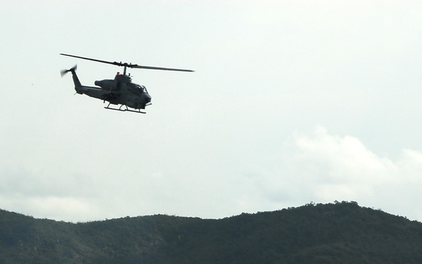 The image depicts an amphibious military assault exercise with multiple armored amphibious vehicles (AAVs) approaching and landing on a beach while a military attack helicopter provides aerial support overhead. Approximately seven to eight amphibious assault vehicles are visible in various stages of the beach landing, transitioning from water to shore. A forested hillside and overcast sky form the backdrop of the scene.
