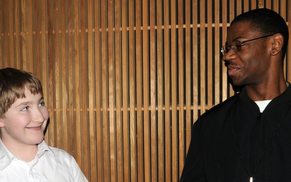 Two individuals are engaged in an interaction where an item appears to be exchanged or presented between them. A young boy in a white shirt is smiling while a taller adult male in a black outfit with a lanyard/badge hands him what appears to be a small booklet or card. The setting appears to be an indoor venue with a distinctive wooden slatted wall in the background.