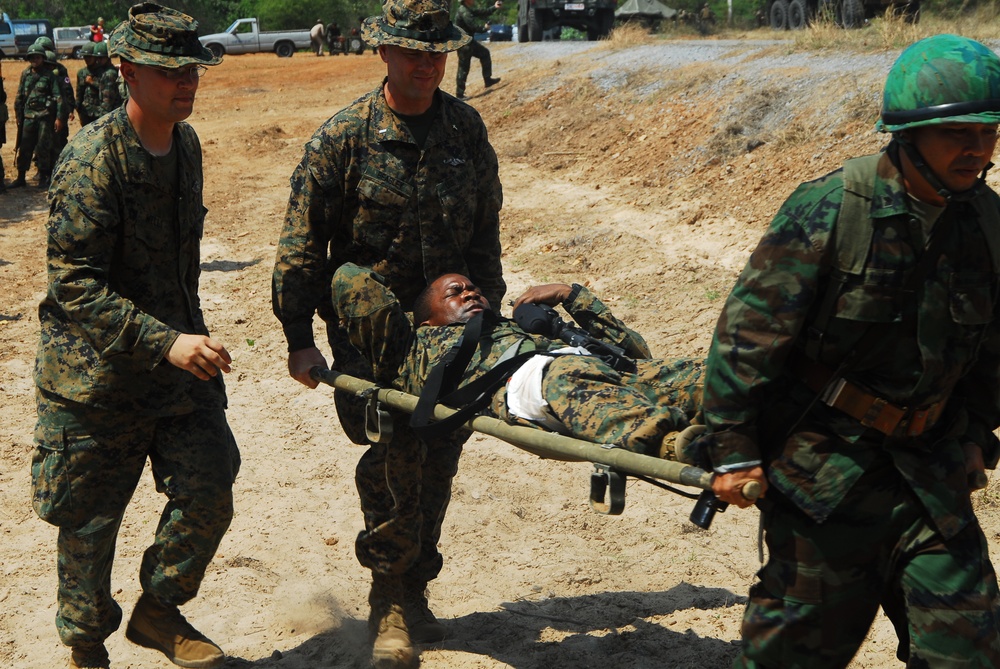 Military personnel in camouflage uniforms are carrying a soldier on a stretcher across sandy terrain. The scene appears to depict a casualty evacuation drill or training exercise involving what appear to be U.S. Marines in MARPAT digital camouflage alongside soldiers in a different camouflage pattern wearing green helmets. Military vehicles and additional personnel are visible in the background.