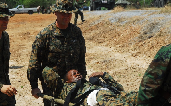 Military personnel in camouflage uniforms are carrying a soldier on a stretcher across sandy terrain. The scene appears to depict a casualty evacuation drill or training exercise involving what appear to be U.S. Marines in MARPAT digital camouflage alongside soldiers in a different camouflage pattern wearing green helmets. Military vehicles and additional personnel are visible in the background.