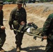 Military personnel in camouflage uniforms are carrying a soldier on a stretcher across sandy terrain. The scene appears to depict a casualty evacuation drill or training exercise involving what appear to be U.S. Marines in MARPAT digital camouflage alongside soldiers in a different camouflage pattern wearing green helmets. Military vehicles and additional personnel are visible in the background.