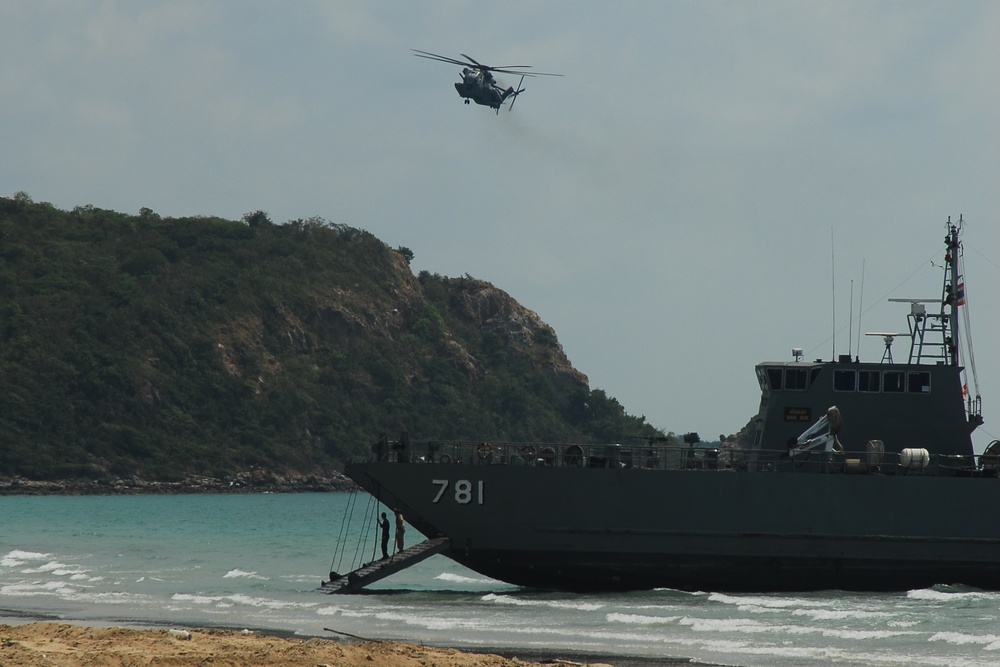 A military landing craft bearing hull number 781 is beached on a sandy shoreline, with its bow ramp lowered toward the beach. A large military helicopter, consistent with a CH-53 Sea Stallion type, flies overhead. The scene appears to depict an amphibious military exercise or operation near a rocky, vegetation-covered coastal headland.