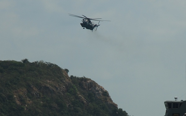 A military landing craft bearing hull number 781 is beached on a sandy shoreline, with its bow ramp lowered toward the beach. A large military helicopter, consistent with a CH-53 Sea Stallion type, flies overhead. The scene appears to depict an amphibious military exercise or operation near a rocky, vegetation-covered coastal headland.