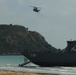 A military landing craft bearing hull number 781 is beached on a sandy shoreline, with its bow ramp lowered toward the beach. A large military helicopter, consistent with a CH-53 Sea Stallion type, flies overhead. The scene appears to depict an amphibious military exercise or operation near a rocky, vegetation-covered coastal headland.
