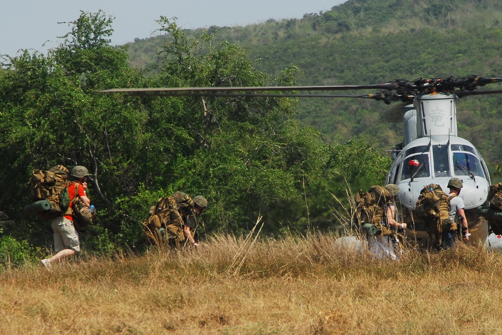 A group of military personnel carrying heavy backpacks and gear are moving toward or away from a light utility helicopter landed in a dry grassy field. At least one civilian in civilian clothing (orange shirt, khaki shorts) is visible among the group. The setting appears to be a rural or semi-arid environment with dense green vegetation and hills in the background.
