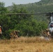A group of military personnel carrying heavy backpacks and gear are moving toward or away from a light utility helicopter landed in a dry grassy field. At least one civilian in civilian clothing (orange shirt, khaki shorts) is visible among the group. The setting appears to be a rural or semi-arid environment with dense green vegetation and hills in the background.