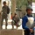 217th Military Police Train Iraqi Police at the Criminal Justice Center The image shows two U.S. Army soldiers in ACU (Army Combat Uniform) camouflage alongside a local security personnel wearing a blue shirt, body armor vest, and green helmet. The local guard is holding an assault rifle and appears to be on active security duty. The setting appears to be a public or government compound in the Middle East, likely Iraq, based on the architecture and signage.