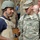 217th Military Police train Iraqi police at the Criminal Justice Center The image shows a U.S. Army Military Police soldier named McClellan in ACU (Army Combat Uniform) conversing with a person wearing a body armor vest and helmet bearing what appears to be an Iraqi police or security force patch. The individual on the left is holding a wooden object, possibly a training prop or tool. Several other personnel are visible in the background in what appears to be an outdoor setting.