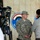 217th Military Police train Iraqi police at the Criminal Justice Center A U.S. Army soldier named 'Worthy' (visible on name tape) wearing ACU digital camouflage uniform stands in front of a wall bearing what appears to be an Iraqi police or security force emblem. A television camera operator is visible in the foreground left, suggesting a media interview or press event is taking place. Several civilians are present, with one appearing to engage the soldier in conversation.