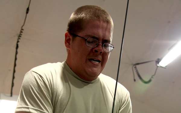 A young male soldier in U.S. Army ACU (Army Combat Uniform) bottoms and a sage green t-shirt is performing a cable pull-down or tricep pushdown exercise on a weight machine inside what appears to be a military tent gym. He is wearing glasses and workout gloves, and his facial expression shows physical exertion. A 'Body Master' brand weight machine is visible in the background.