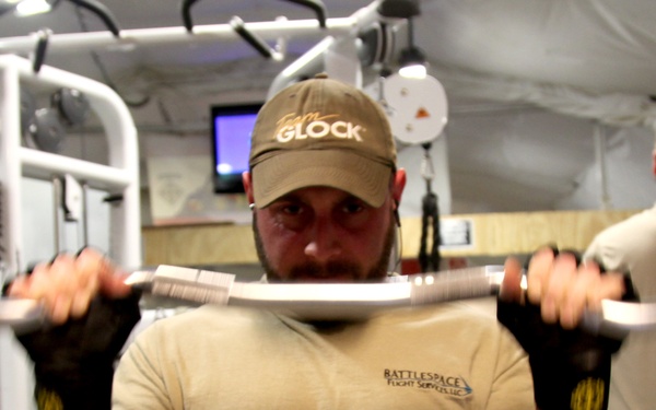 A muscular man wearing a 'GLOCK' cap and tan military-style shirt is performing a barbell curl exercise in what appears to be a military forward operating base (FOB) gym. The gym is housed in a tent-like structure with white fabric walls and wooden paneling. A second person in military camouflage pants is visible in the background using other equipment.